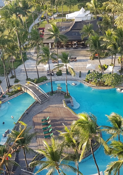 An aerial view captures a luxurious resort area featuring a large swimming pool surrounded by palm trees. The pool has a unique shape with a central island connected by small bridges. Sun loungers with umbrellas line the poolside. To the left, a lawn overlooks a body of water, with a lighthouse structure visible. The entire setting exudes a tropical and serene atmosphere.