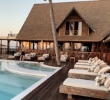 A luxurious wooden resort with a thatched roof, featuring a pool surrounded by lounge chairs and a wooden deck. There are a few people relaxing and walking around the area, and the setting sun casts a warm glow on the building and surroundings.
