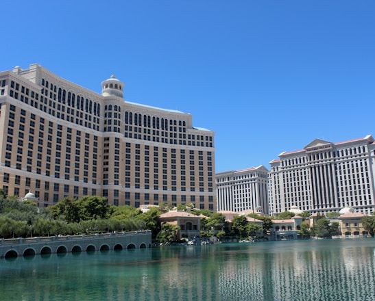 A large luxury hotel and casino building with multiple stories, featuring a light facade with arched windows. The structure has a prominent central tower with a dome-like top. It is surrounded by landscaping with trees and a body of water in the foreground.