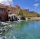 A tropical resort scene featuring a large, pink-colored hotel with spires, surrounded by palm trees and lush greenery. A serene, crystal-clear turquoise pond is in the foreground, accented by small waterfalls cascading from rocks. The sky is bright blue with large, fluffy white clouds.