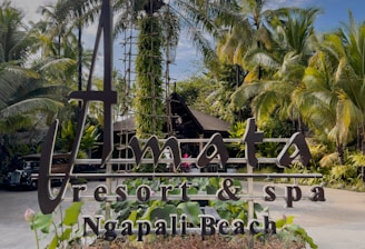 A resort sign is prominently displayed, surrounded by lush tropical plants and palm trees. The sky is blue with scattered clouds, creating a serene atmosphere.