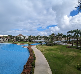 A luxurious resort setting featuring a large, winding swimming pool surrounded by manicured lawns and lush tropical palm trees. There are thatched-roof structures and white buildings in the background, with a partly cloudy sky overhead.