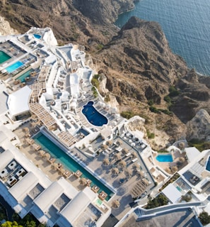 An aerial view of a luxurious coastal resort with multiple swimming pools. The resort features white buildings with terraces and sun loungers, nestled into rocky cliffs overlooking a blue ocean. The landscape is a blend of architectural elegance and natural beauty.